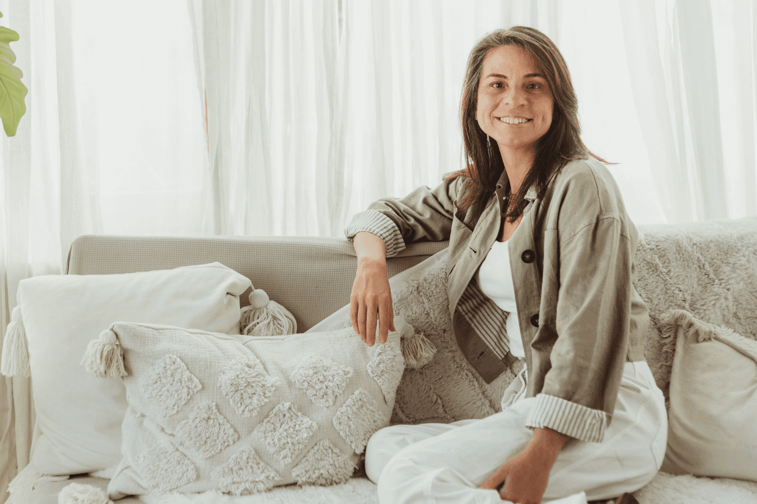 Woman sitting on a cozy couch with decorative pillows and soft lighting, smiling at the camera.